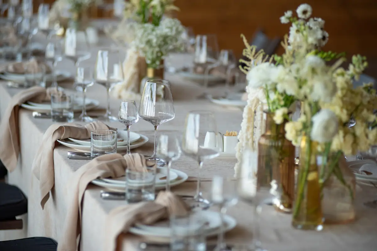 Reception Table Styled With Soft Neutral Linens, Layered Glassware, And Floral Centerpieces Designed To Create Depth Under Indoor Lighting