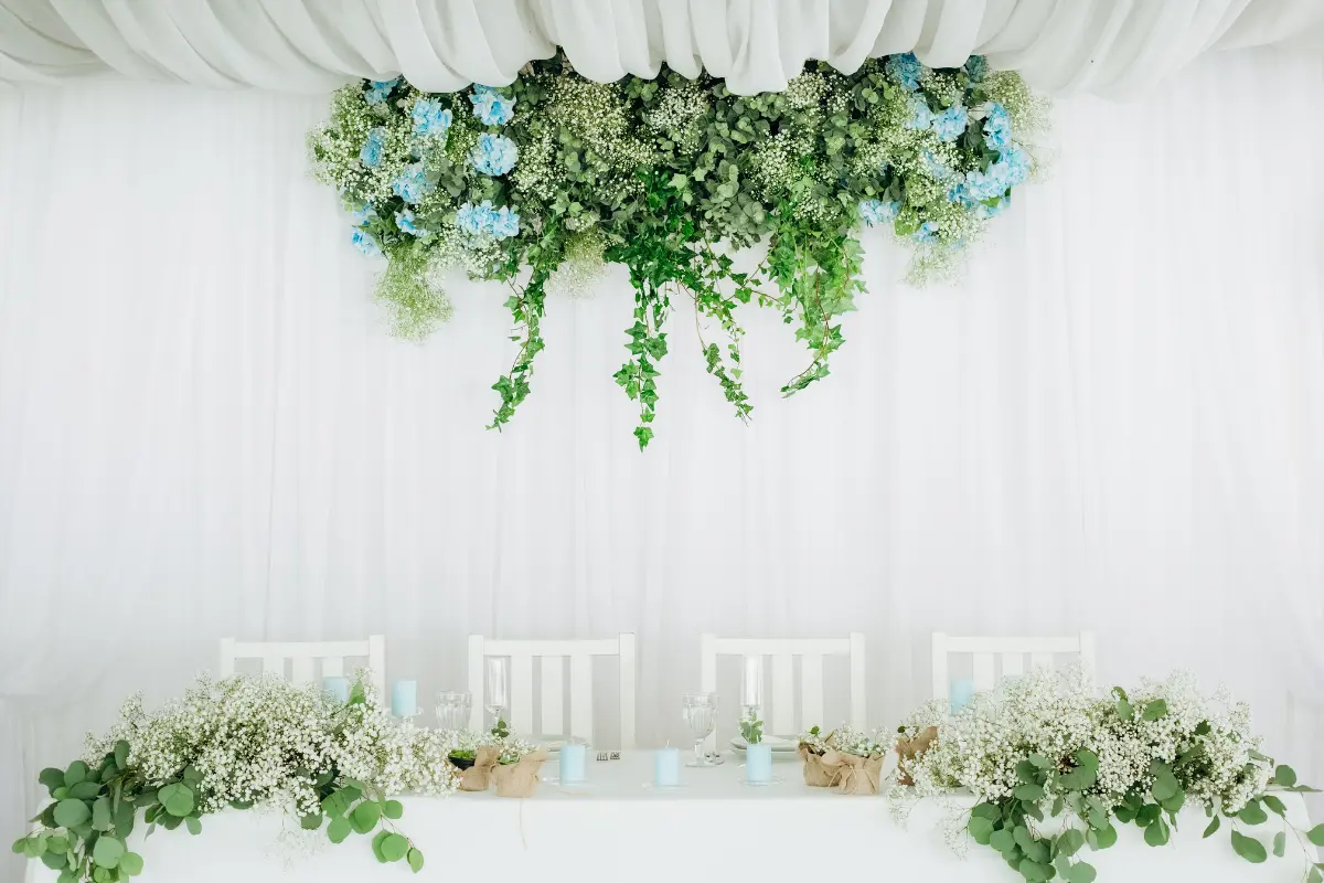 White-Draped Head Table Framed With Cascading Greenery And Soft Blue Florals, Creating A Balanced And Immersive Reception Focal Point